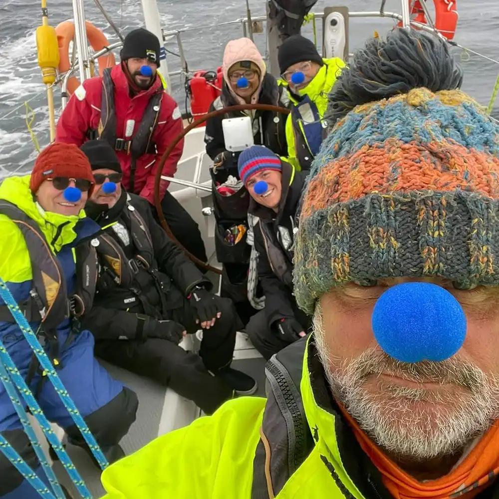 A group of sailors on deck during an Arctic expedition, all wearing life jackets and blue noses in celebration of crossing the Arctic Circle and earning the Order of the Blue Nose. Dressed in warm sailing gear, they smile against a backdrop of open ocean and overcast skies, marking a milestone in their high-latitude adventure.