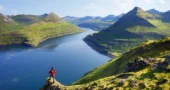 A hiker in a red jacket stands on a grassy cliff overlooking a deep fjord in the Faroe Islands, surrounded by towering, rugged mountains. Below, the winding waterway leads to the open sea, a dramatic route for sailors exploring this remote North Atlantic archipelago. The scene captures the wild, untouched beauty of the Faroes—an unforgettable destination for an offshore sailing adventure.