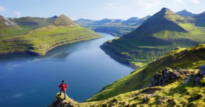 A hiker in a red jacket stands on a grassy cliff overlooking a deep fjord in the Faroe Islands, surrounded by towering, rugged mountains. Below, the winding waterway leads to the open sea, a dramatic route for sailors exploring this remote North Atlantic archipelago. The scene captures the wild, untouched beauty of the Faroes—an unforgettable destination for an offshore sailing adventure.