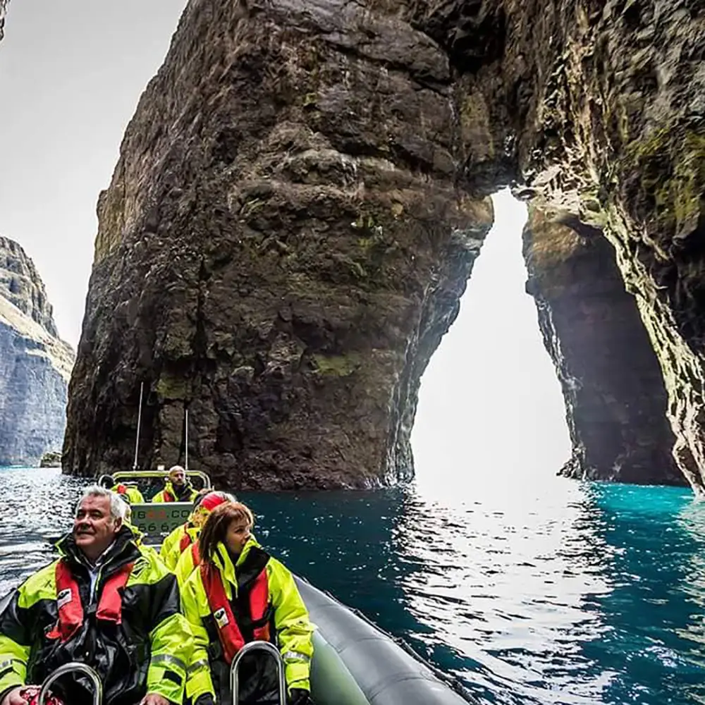 A group of people wearing bright yellow waterproof jackets and life vests sit in a rigid inflatable boat (RIB) as they explore a dramatic sea grotto in the Faroe Islands. Towering dark rock formations rise on either side, with a natural archway framing the bright open ocean beyond. The water is a deep blue, reflecting light from the cave entrance. The passengers gaze in awe at the rugged beauty surrounding them.