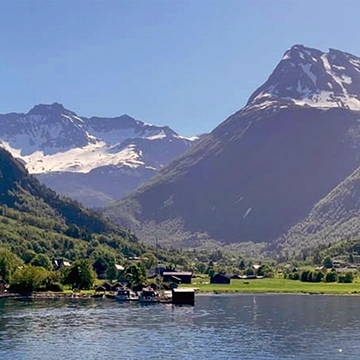 A peaceful Norwegian fjord-side village, nestled beneath towering snow-capped mountains. The still waters reflect the surrounding peaks, capturing the raw beauty of Norway’s west coast.