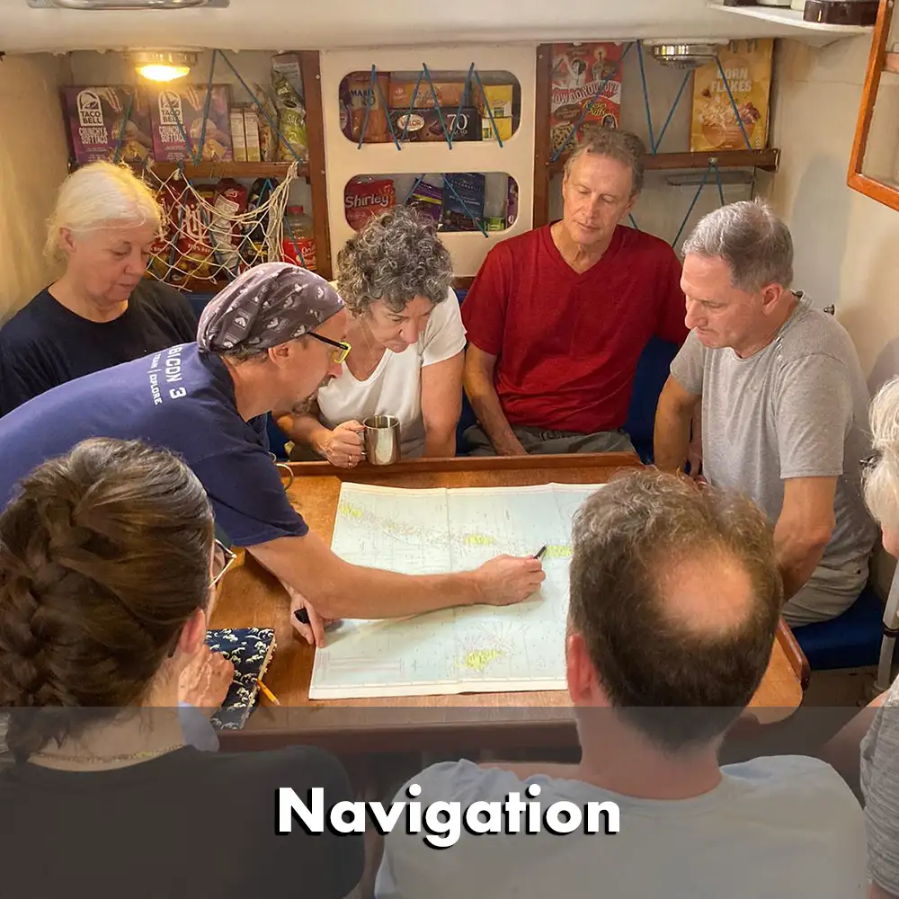 Équipage réuni autour d'une table de navigation dans le salon d'un yacht, examinant des cartes marines. Une personne pointe un endroit sur la carte tandis que d'autres observent attentivement et discutent. Les étagères à l'arrière-plan sont remplies de nourriture et de fournitures.  