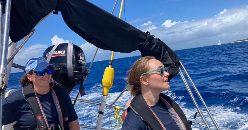 Two female sailors steering a yacht during the 2025 St. Maarten Heineken Regatta. The woman at the helm, wearing light blue sunglasses and a navy sailing shirt, focuses ahead while gripping the wheel. Beside her, another crew member in a blue cap, sunglasses, and a life jacket smiles while monitoring the race. The deep blue ocean and a distant sailboat are visible under a bright sky, capturing the excitement of offshore racing.