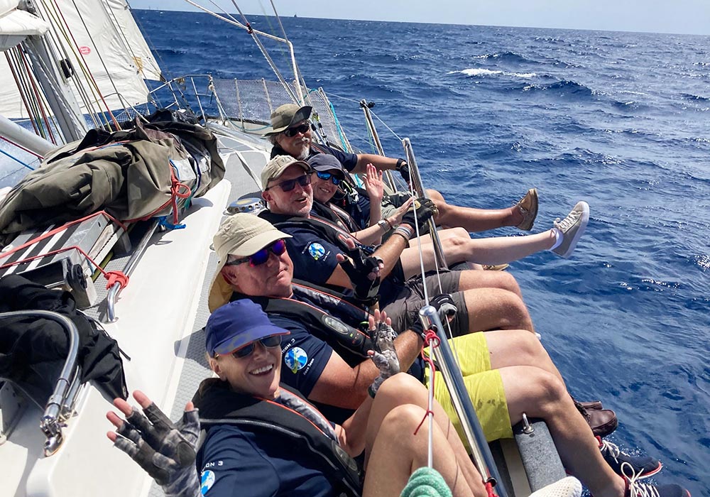 Eine Gruppe von Seglern wandert auf der Reling einer Yacht während der St. Maarten Heineken Regatta 2025. Bekleidet mit marineblauen Segelhemden, Schwimmwesten und Sonnenbrillen lehnt sich die Crew über die Bordwand und balanciert das Boot aus, während sie lächelt und in die Kamera winkt. Der tiefblaue Ozean erstreckt sich bis zum Horizont, die Segel sind getrimmt und die Leinen gespannt, um die Aufregung und das Teamwork der Hochseeregatten einzufangen.  