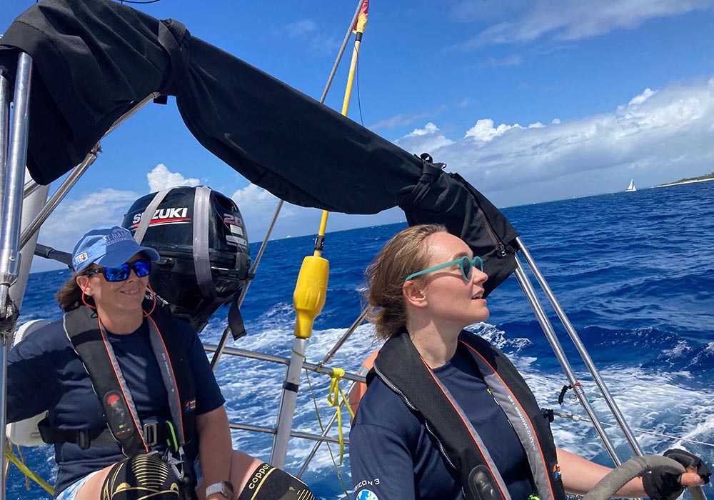 Two female sailors steering a yacht during the 2025 St. Maarten Heineken Regatta. The woman at the helm, wearing light blue sunglasses and a navy sailing shirt, focuses ahead while gripping the wheel. Beside her, another crew member in a blue cap, sunglasses, and a life jacket smiles while monitoring the race. The deep blue ocean and a distant sailboat are visible under a bright sky, capturing the excitement of offshore racing.